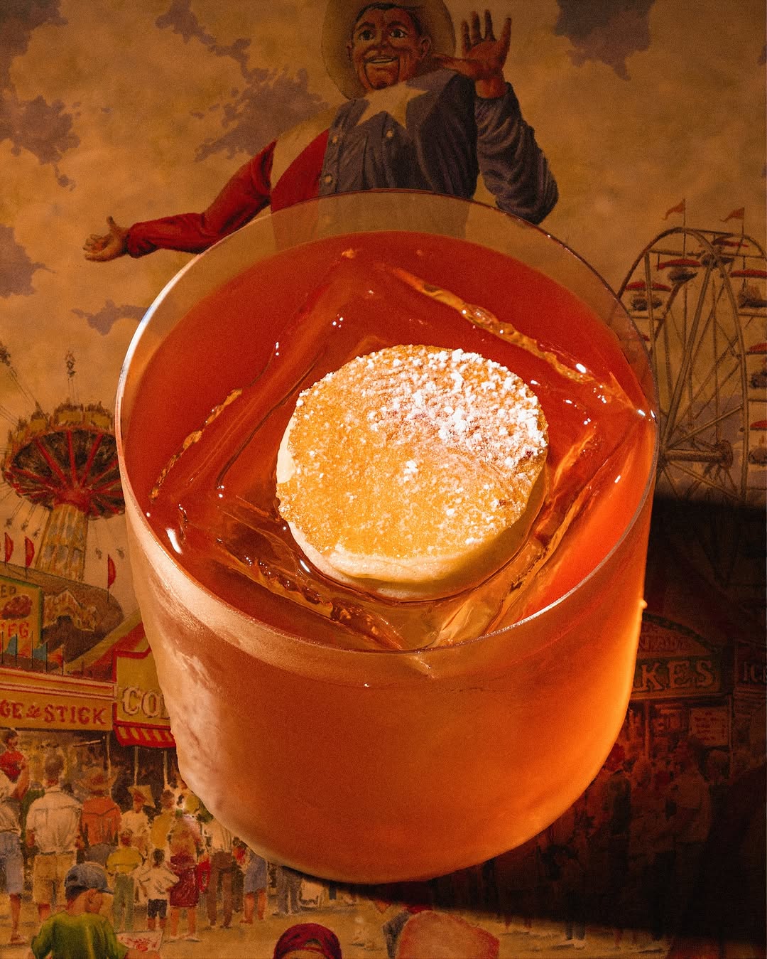 Amber cocktail over a single large ice cube, set against a painted state fair mural